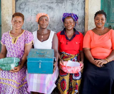 Four women sit together. Two are holding baskets with papers and one is holding a metal box.